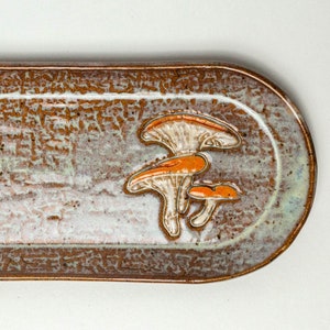 Puede incluir: Bandeja de cerámica ovalada con superficie moteada marrón y blanca y borde elevado. La bandeja presenta un grupo de tres setas naranjas y blancas como elemento decorativo. Ideal para servir alimentos.