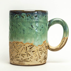 May include: A green and brown ceramic mug with a dragonfly design. The mug has a textured surface and a handle.