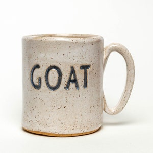 May include: A white ceramic mug with a speckled glaze and a black handle. The word "GOAT" is written in black on the front of the mug.