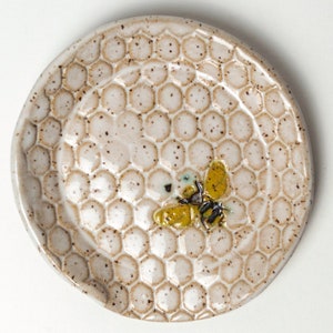 Op de afbeelding: Een klein, rond keramisch schaaltje met een honingraatpatroon en een geel en zwart bijenontwerp. Het schaaltje is gebroken wit met bruine spikkels en een opstaande rand. De bij heeft gele vleugels en zwarte strepen.