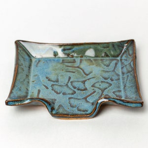 May include: A rectangular ceramic dish with a blue and green glaze. The dish has a unique shape with a curved edge and a raised rim. The surface features a leaf pattern and a bronze-colored trim.
