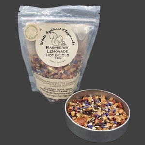 May include: A clear plastic bag and a small metal tin filled with loose leaf tea. The bag has a label that reads "Raspberry Lemonade Hot & Cold Tea." The tea blend includes dried fruits and blue flower petals. The package is 2 oz (56.6g).