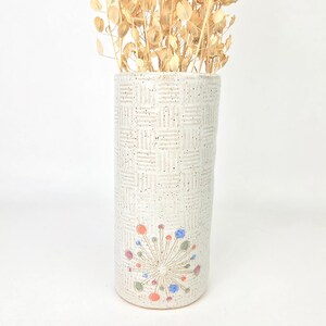 May include: A tall, off-white ceramic vase with a textured, woven pattern. The vase features a colorful, starburst design near the base and is filled with dried, tan-colored plant stems. The vase is approximately 10 inches tall.