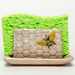 May include: A ceramic sponge holder with a honeycomb design and a bee illustration. The holder has a small tray for catching water and is designed to hold a green sponge.