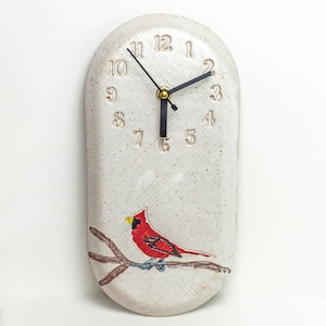 May include: Oval ceramic wall clock with a speckled, off-white background. The clock has black hands, gold accents, and embossed numbers. A red cardinal bird with a yellow beak is perched on a brown branch at the bottom.