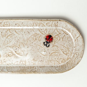 Könnte beinhalten: Eine weiße Keramik-Servierplatte mit einem erhabenen Blatt-Muster und einem kleinen roten Marienkäfer-Design.