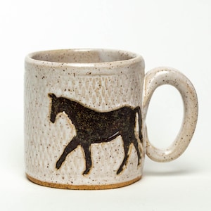 Puede incluir: Una taza de cerámica blanca con una silueta marrón de un caballo en la parte delantera. La taza tiene un esmalte moteado y un asa marrón.