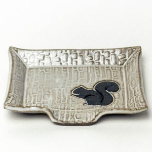 May include: A white ceramic dish with a textured surface and a black squirrel design. The dish has a slightly curved edge and a small indentation on one side.