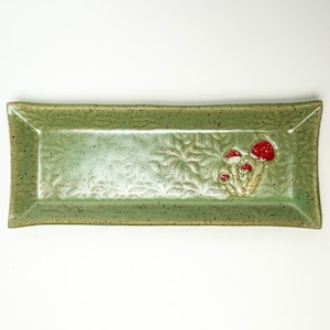 Puede incluir: Bandeja rectangular de cerámica en un esmalte verde moteado. La bandeja presenta un patrón de hojas en relieve y un grupo de setas rojas y beige en un extremo. Ideal para servir aperitivos o exhibir objetos pequeños.