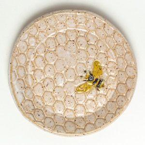May include: A ceramic dish with a honeycomb pattern and a yellow bee design. The dish is white with brown speckles.