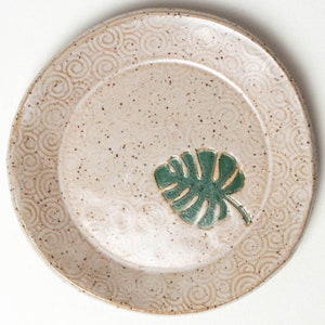 May include: A speckled ceramic plate with a light beige background and a teal green monstera leaf design. The plate's rim features a repeating spiral pattern in a darker beige color. The plate is a handmade item.
