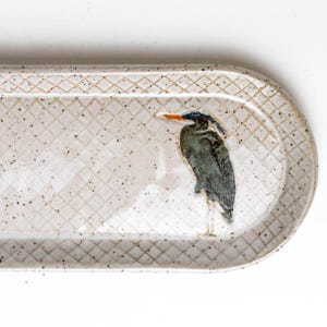 May include: A ceramic serving platter with a heron design. The oval-shaped platter has a cream-colored background with a diamond pattern and speckled details. The heron is depicted in shades of gray, blue, and orange.