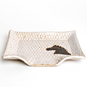 May include: A rectangular, off-white ceramic dish with a textured surface and a dark brown horse silhouette. The dish has a slightly curved design on the bottom edge and is speckled with small, dark brown spots.