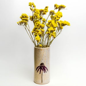 May include: A ceramic vase with a speckled glaze and a purple flower design. The vase is filled with a bouquet of dried yellow flowers.