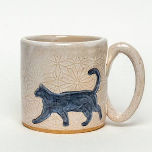 Puede incluir: Taza de cerámica beige con una silueta de gato azul oscuro. La taza presenta un patrón geométrico texturizado y un asa curva. El gato camina, con la cola curvada hacia arriba.