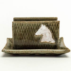 May include: A ceramic business card holder with a horse head design. The holder is a muted green with a white horse head silhouette. The base is a matching green, and the holder has a textured pattern.