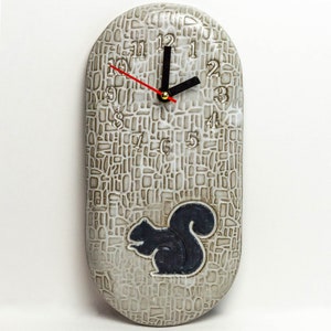 May include: A white ceramic oval clock with a black squirrel silhouette and black clock hands on a white background with a textured pattern. The clock face has black numbers 1 through 12.