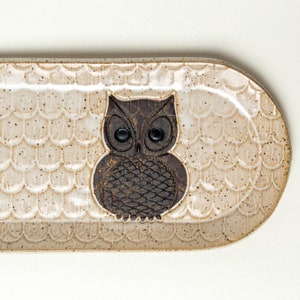 May include: A ceramic oval serving dish with a brown owl design. The dish has a speckled beige glaze and a raised rim.