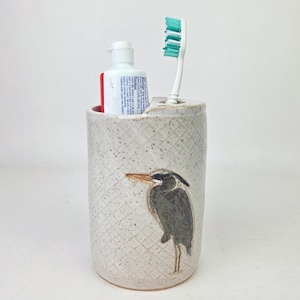 May include: A speckled ceramic toothbrush holder with a heron illustration. The holder contains a tube of toothpaste and a toothbrush with a green bristle head. The holder has a diamond pattern and is a neutral color.