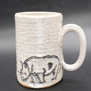 Könnte beinhalten: Cremefarbene Keramiktasse mit einer strukturierten, ziegelartigen Oberfläche und einem großen Henkel. Auf der Vorderseite befindet sich eine schwarz-weiße Nashorn-Illustration. Geeignet für Kaffee, Tee oder andere Getränke. Höhe ca. 12 cm.