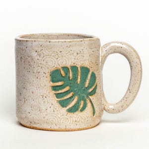 Puede incluir: Una taza de cerámica blanca con un diseño de hoja verde. La taza tiene un esmalte moteado y una superficie texturizada.