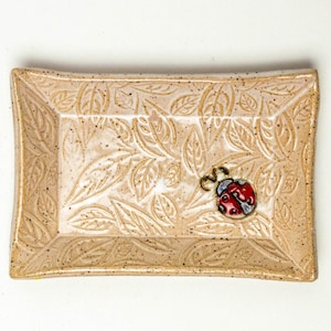 May include: A rectangular ceramic dish with a raised leaf pattern and a small ladybug design. The dish is beige with brown speckles.