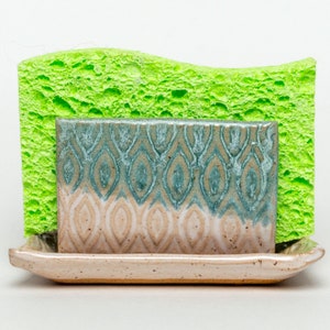 May include: A ceramic sponge holder with a light brown glaze and a blue and white geometric pattern. The holder has a rectangular shape and a raised lip around the edge. A bright green sponge is sitting on top of the holder.