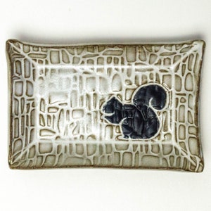 May include: A rectangular ceramic dish with a textured surface and a black silhouette of a squirrel in the center.