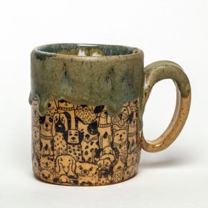 Könnte beinhalten: Keramikbecher mit einem einzigartigen Design mit einer Vielzahl von schwarz-weißen Hundeillustrationen. Der obere Rand hat eine grüne Glasur, die an der Seite heruntertropft. Der Griff und der Boden sind hellbraun.