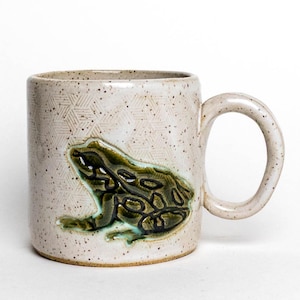 Pode incluir: Uma caneca de cerâmica salpicada com um padrão geométrico texturizado e um design de rã esculpida. A rã é verde e castanha com detalhes em azul. A caneca tem uma pega arredondada e uma paleta de cores neutras.