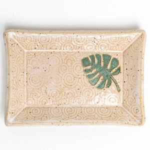 May include: A rectangular ceramic dish with a white glaze and a brown speckled pattern. The dish has a raised border and a green leaf design in the center.