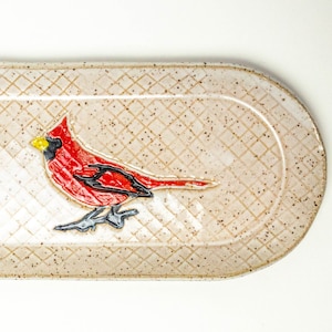 Puede incluir: Una bandeja de servir ovalada de cerámica con fondo beige y un diseño de pájaro cardenal rojo. La bandeja tiene una superficie texturizada con un patrón de diamante.