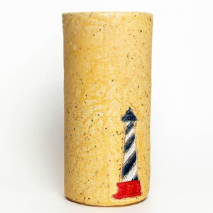 May include: A cylindrical ceramic vase with a textured, light yellow surface. A lighthouse design is painted on the front, featuring black and white stripes, a blue top, and a red base. The vase has a smooth, rounded top.
