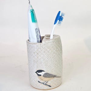 Könnte beinhalten: Ein gesprenkelter Zahnbürstenhalter aus Keramik mit einem Vogeldesign. Der Halter enthält eine Tube Zahnpasta und eine Zahnbürste mit blauem Griff. Der Halter hat ein Rautenmuster und ist cremefarben.