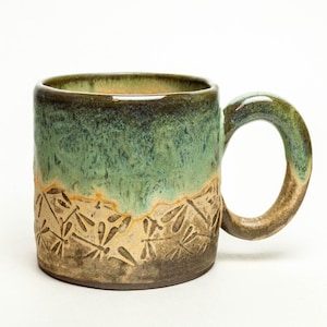 May include: A green and brown ceramic mug with a dragonfly design. The mug has a wide, rounded handle.