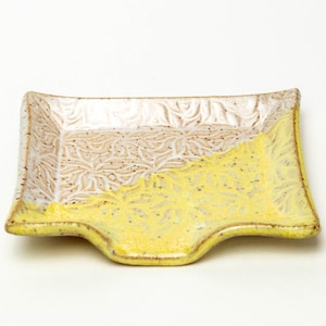 Op de afbeelding: Een klein, vierkant keramisch schaaltje met een gele glazuur op de onderste helft en een witte glazuur op de bovenste helft. Het schaaltje heeft een decoratief bladmotief op beide helften.