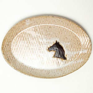 Puede incluir: Un plato ovalado de cerámica blanca con un diseño de silueta de caballo marrón. El plato tiene un esmalte moteado y una superficie texturizada.