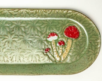 May include: A green ceramic serving dish with a raised design of red and white mushrooms. The dish has a speckled glaze and a slightly oval shape.