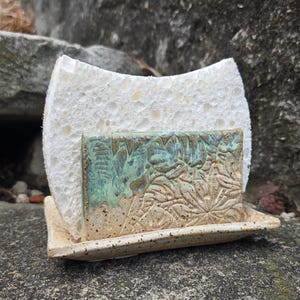 May include: A ceramic sponge holder with a rectangular base and a vertical front panel featuring a textured floral pattern in shades of blue and beige. The holder contains a white, porous, rectangular sponge that rests against the front panel.