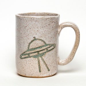 May include: A white ceramic mug with a speckled glaze and a green UFO design.