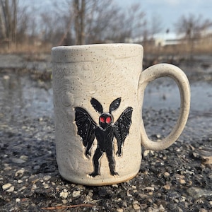 May include: A speckled off-white ceramic mug featuring a raised, black relief of a winged creature with prominent ears and glowing red eyes. The mug has a cylindrical body, a sturdy handle, and a textured surface with subtle moon-like craters.