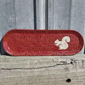 May include: An elongated oval ceramic tray with a deep red glaze and a textured surface pattern of small rectangular shapes. A raised, off-white silhouette of a squirrel is positioned on the right side of the tray, contrasting with the red.