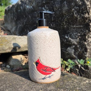 May include: A cylindrical ceramic soap dispenser with a speckled off-white glaze and a textured diamond pattern. The front features a hand-painted red cardinal bird with black accents. The dispenser is topped with a black plastic pump mechanism.