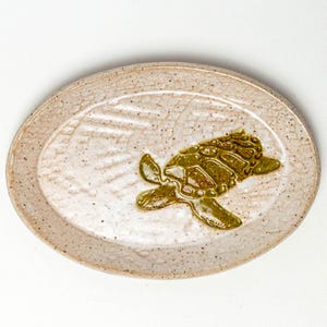 May include: Oval ceramic serving platter with a textured, speckled surface. The platter features a raised, olive-green turtle design in the center. The rim is a darker shade of beige. A decorative piece for serving.