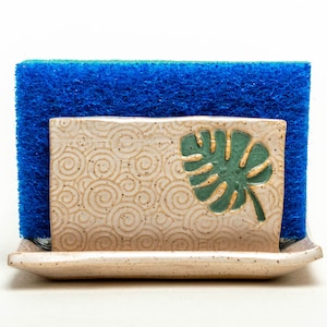 May include: A ceramic sponge holder with a swirling pattern and a green leaf design. The holder has a small tray at the bottom and is designed to hold a blue sponge.
