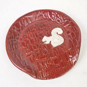 Op de afbeelding: Een handgemaakte, ronde keramische schaal met een textuur, baksteenachtig patroon in een diepe roodbruine kleur. Een wit eekhoorntje is gecentreerd op de schaal. Ideaal voor het serveren van snacks of als decoratief stuk.