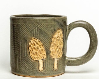 Puede incluir: Una taza de cerámica verde con un patrón geométrico y dos diseños de hongos en relieve. La taza tiene un asa y es perfecta para disfrutar de una bebida caliente.