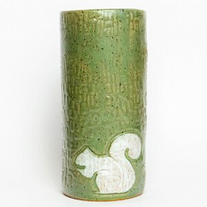 May include: A green ceramic vase with a textured surface and a white squirrel design. The cylindrical vase is approximately 8 inches tall and has a speckled finish. The squirrel is in a side profile.