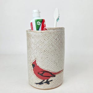 May include: A ceramic toothbrush holder featuring a red cardinal design. The beige holder has a diamond pattern and holds a tube of Colgate toothpaste and a white toothbrush.