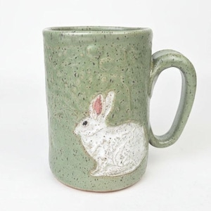 May include: A handmade ceramic mug in a speckled sage green. A white rabbit with pink ears is embossed on the front. The mug has a large handle and a cylindrical shape, perfect for coffee or tea.